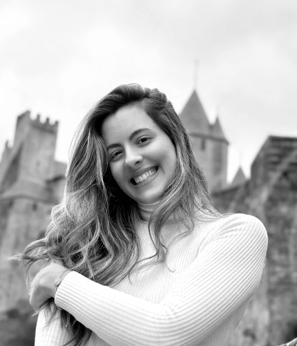 Jahde is a white girl, with long wavy brown hair with blonde highlights, wears a white long-sleeved blouse with a high collar, is smiling, holding her right arm with her left hand, in front of a medieval castle. The photo is with black and white filter.